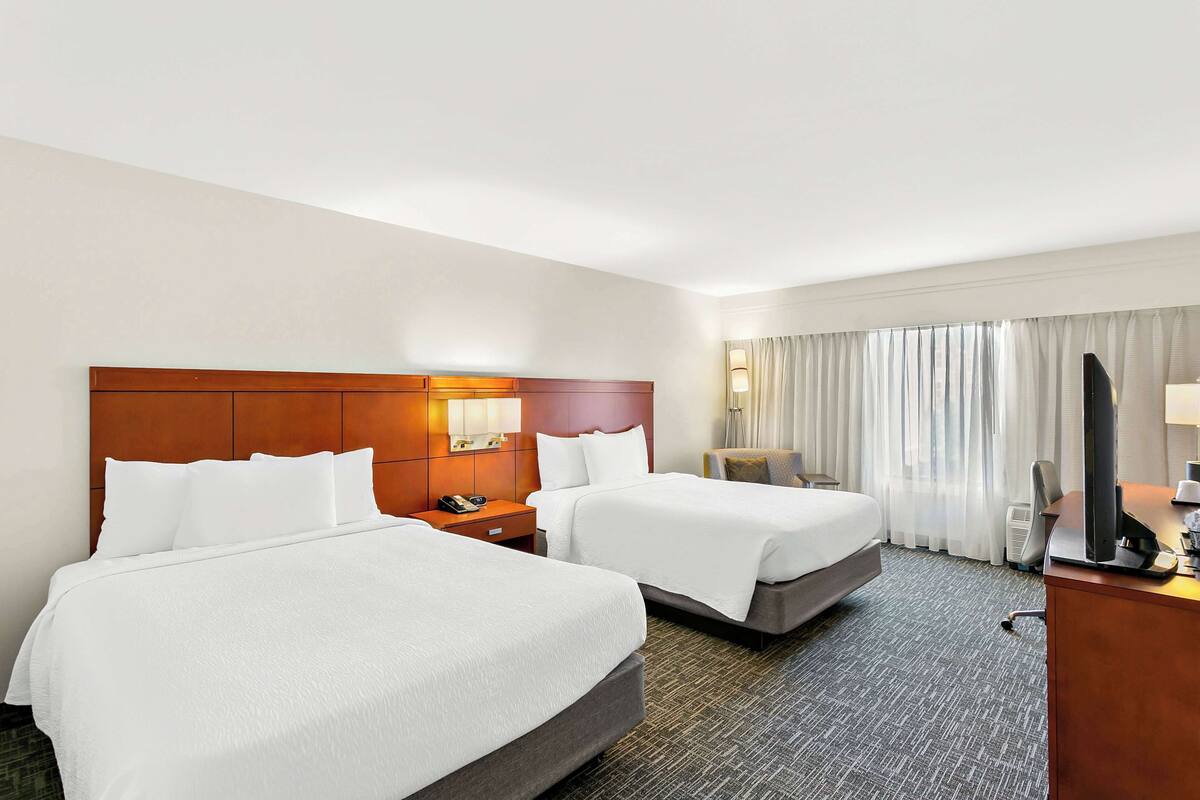 Ein Hotelzimmer mit zwei Betten, einem Holzkopfende, einem Schreibtisch mit Computer und einem großen Fenster mit Vorhängen.