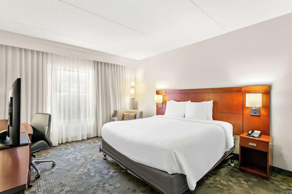 Habitación de hotel con una cama grande, un escritorio con televisor, una silla y una mesita de noche con teléfono.