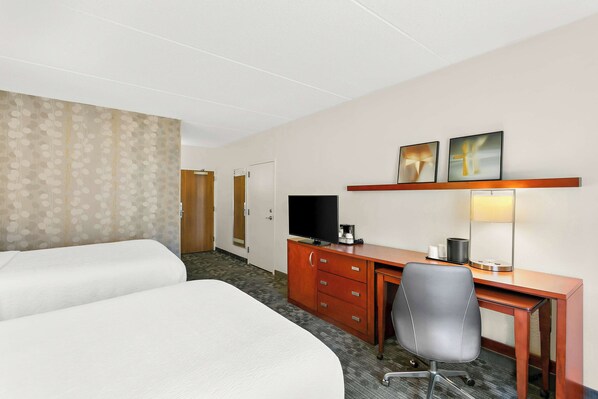 Habitación de hotel con dos camas, un escritorio con silla, televisor, lámpara y puerta que da a otra habitación.