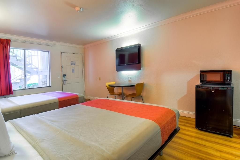 A hotel room with two beds, a TV mounted on the wall, a small table with chairs, and a refrigerator.