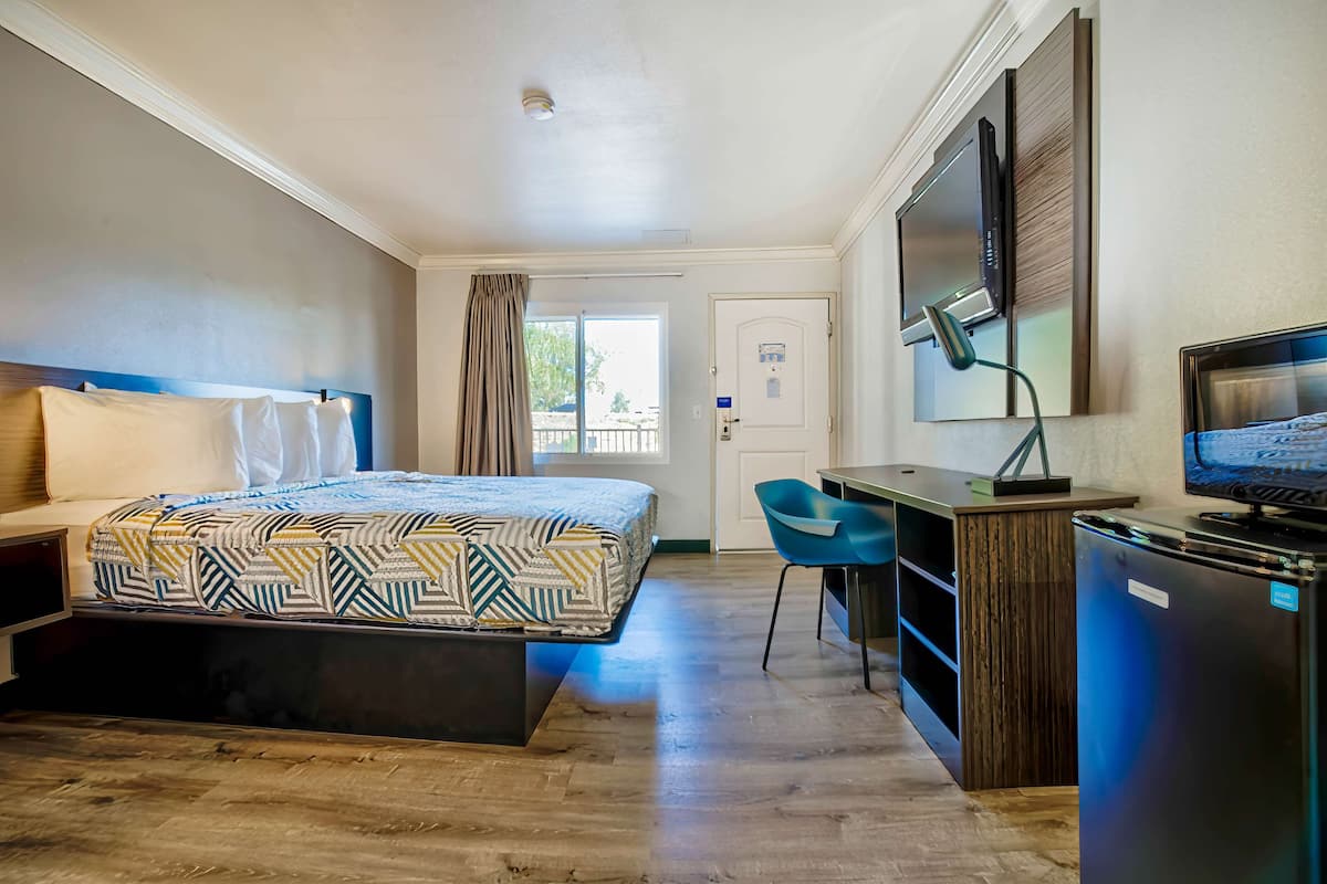 Quarto de hotel com cama, escrivaninha, cadeira, televisão e porta com janela.