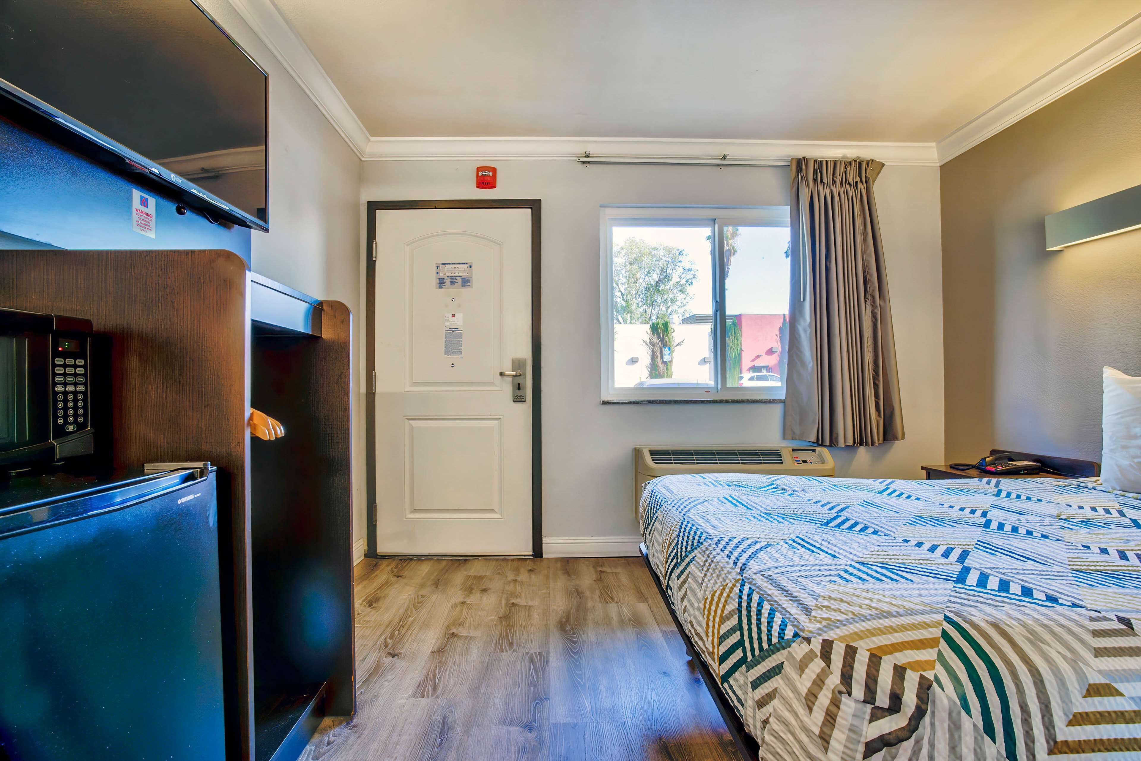 A hotel room with a bed, a microwave, a refrigerator, a door with a lock, a window with curtains, and a thermostat on the wall.
