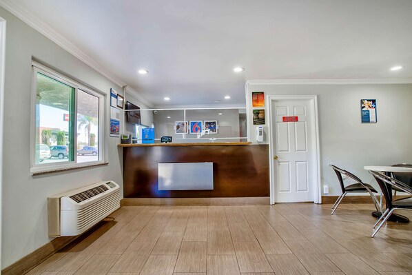 A hotel reception area with a wooden reception desk, a white door, and a window overlooking the street. - Motel 6 Menifee, CA (Menifee)