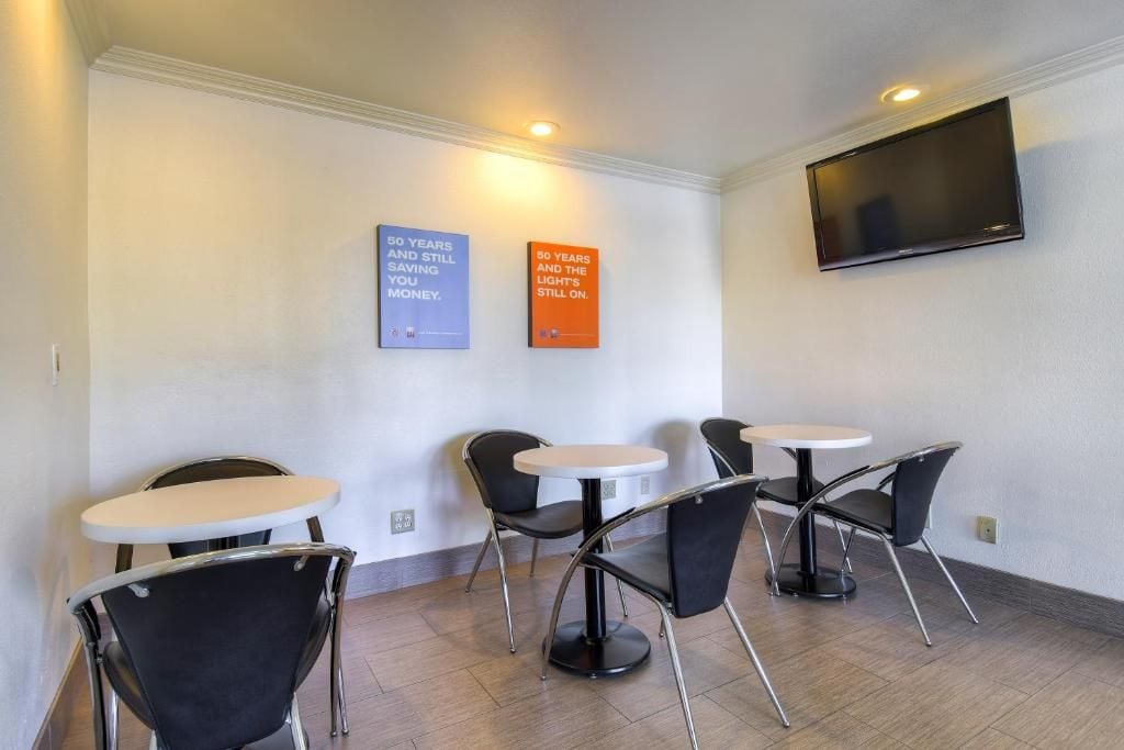 Habitación de hotel con una televisión de pantalla plana montada en la pared, una zona de estar con mesas y sillas redondas, y dos carteles motivacionales en la pared.
