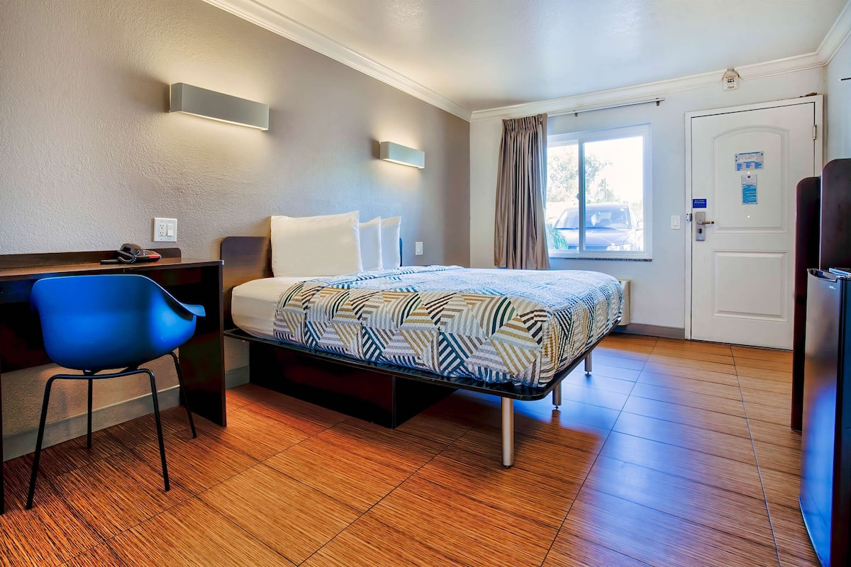 Quarto de hotel com cama, secretária, cadeira e janela com cortinas.