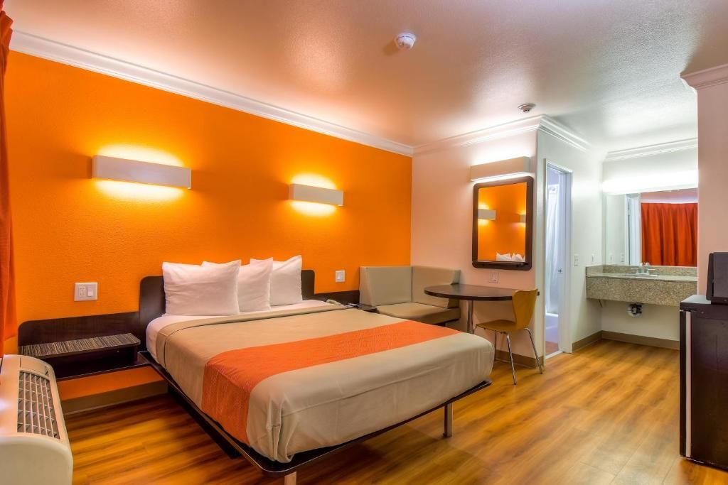 A hotel room with a bed, a desk, a chair, a small table, a mirror, and a kitchenette.