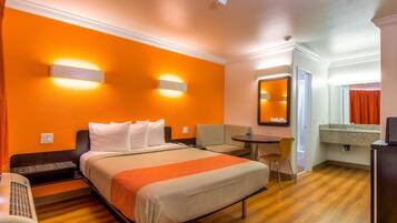 A hotel room with a bed, a desk, a chair, a small table, a mirror, and a kitchenette.