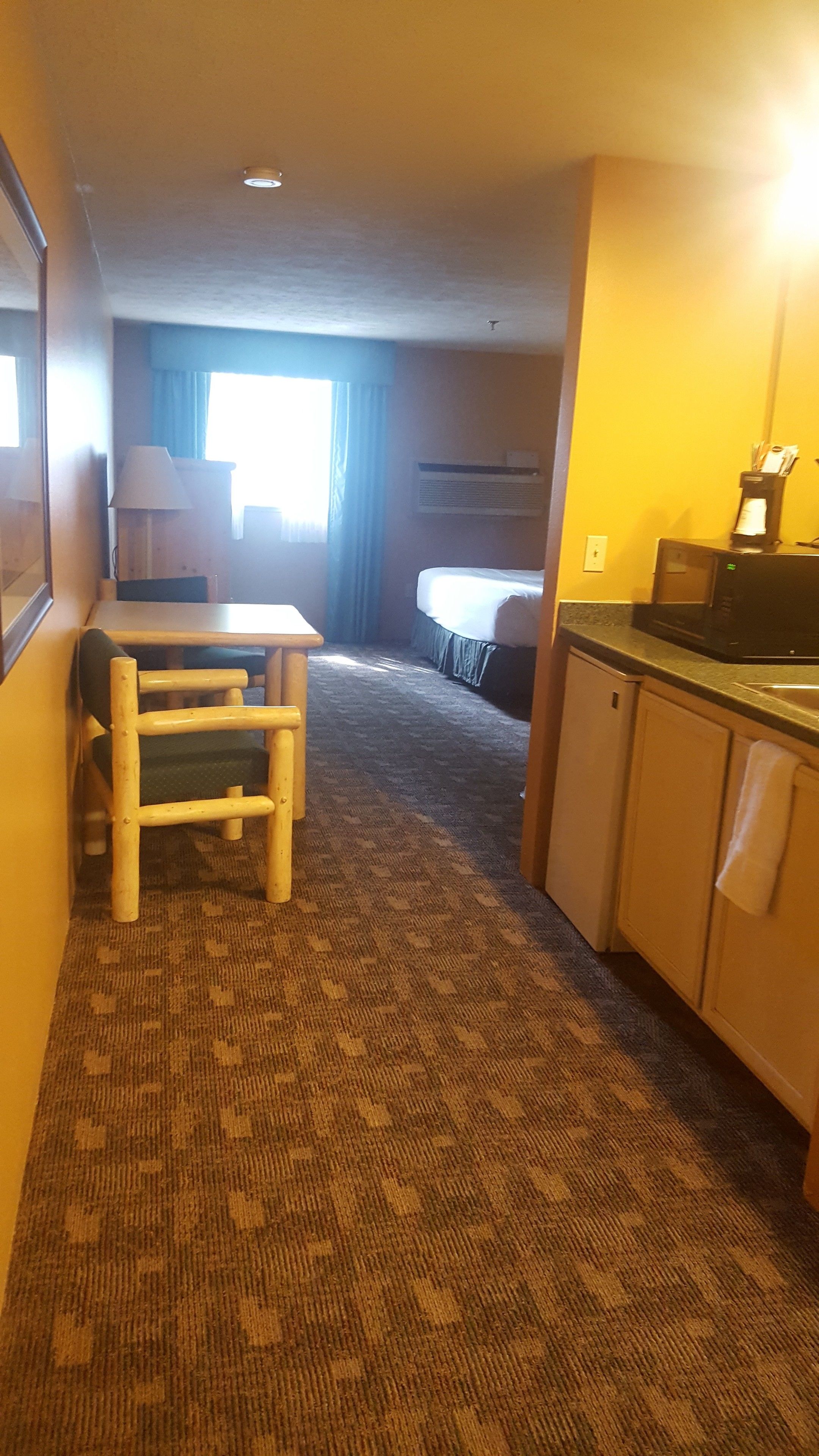 A hotel room with a bed, a desk, a chair, a kitchenette, and a window with curtains.