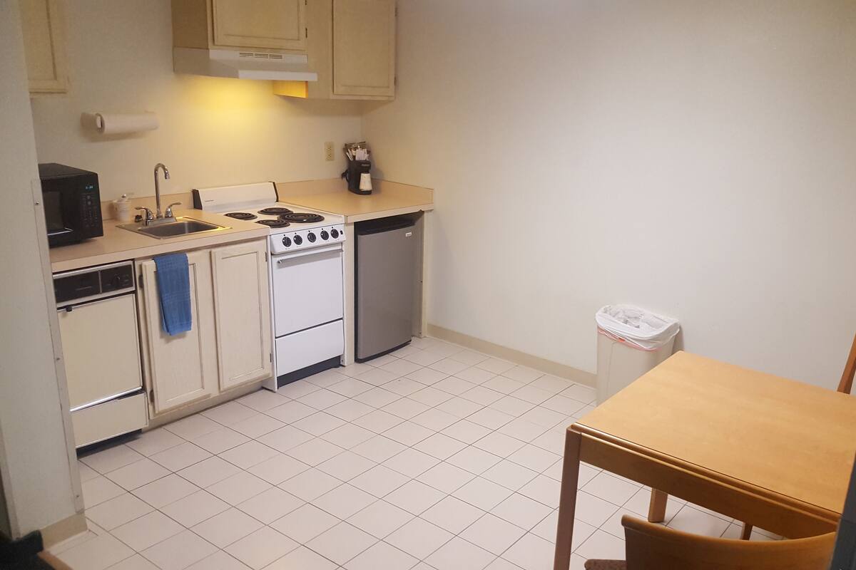 A compact kitchen with white cabinets, a stove, a sink, and a dishwasher. There's a wooden table and a chair in the corner.
