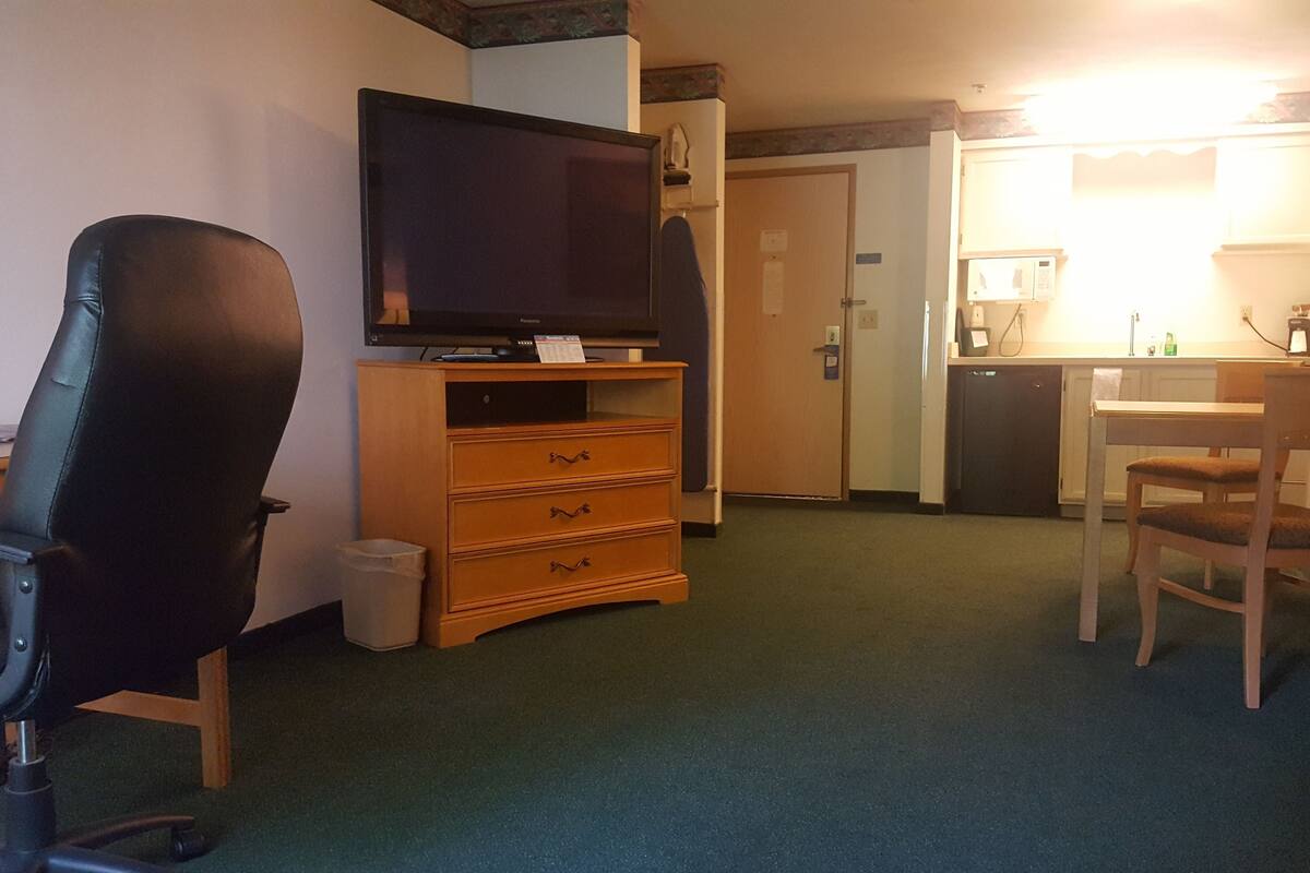 A hotel room with a black office chair, a wooden dresser, a television on a stand, a desk with a chair, and a microwave.