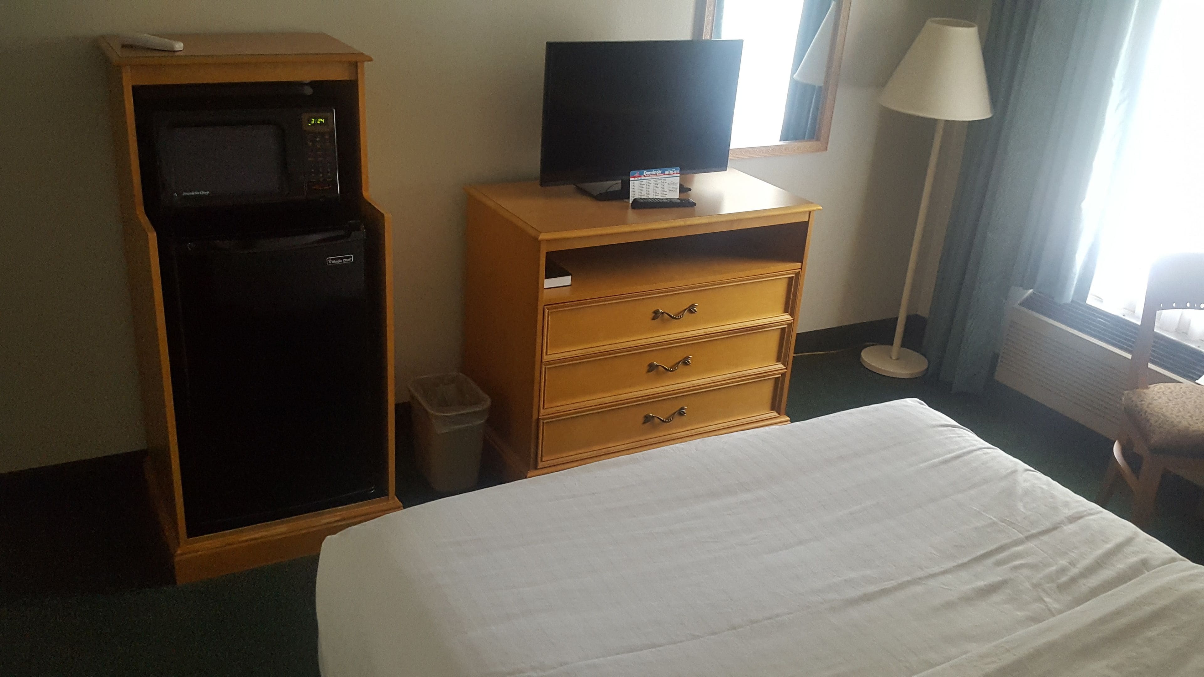 A hotel room with a bed, a television on a wooden stand, a microwave, and a trash bin.