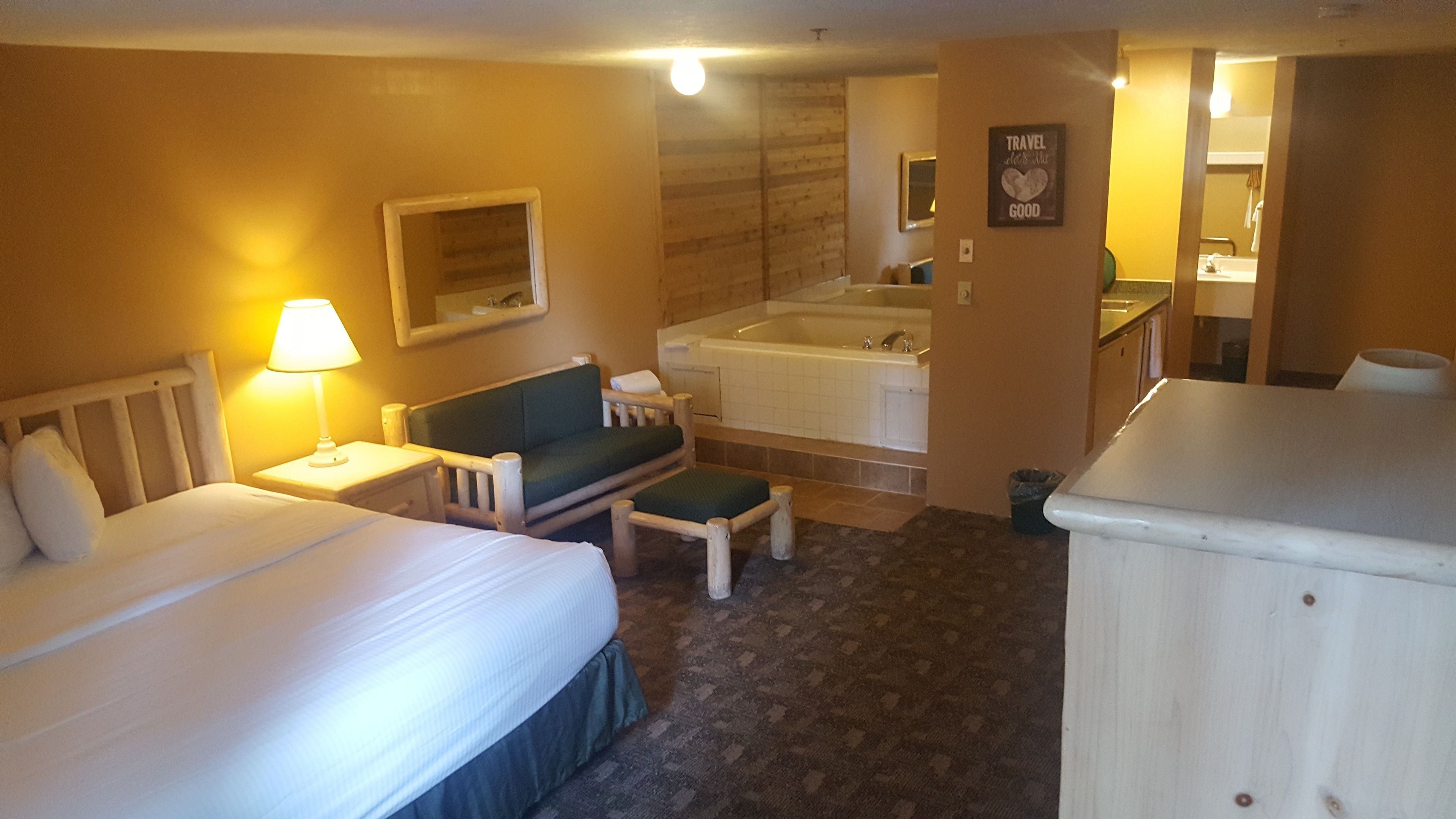 A hotel room with a bed, a small sofa, a chair, a desk, a lamp, a mirror, a bathtub, and a trash bin.