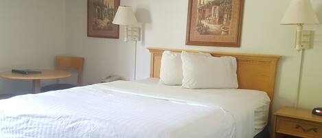 A hotel room with a bed, two paintings on the wall, a round table, a chair, and a nightstand with a lamp.