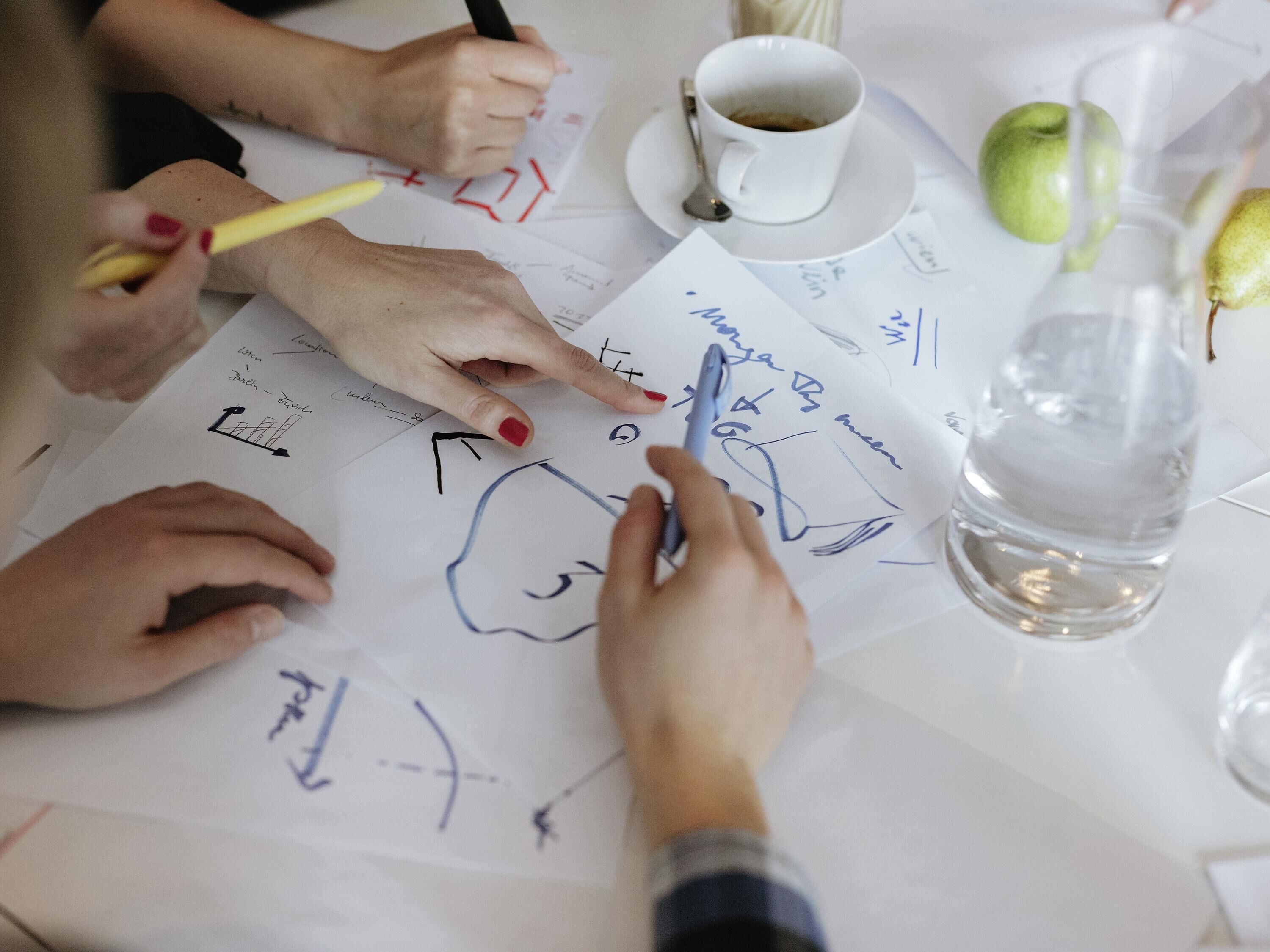 Iemand tekent met een pen op papier, met een koffiekop en een waterfles op tafel.