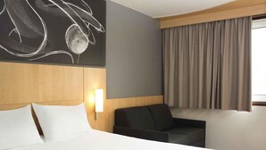 Habitación de hotel con una cama grande, una silla, una lámpara de pared y un ventanal con cortinas.