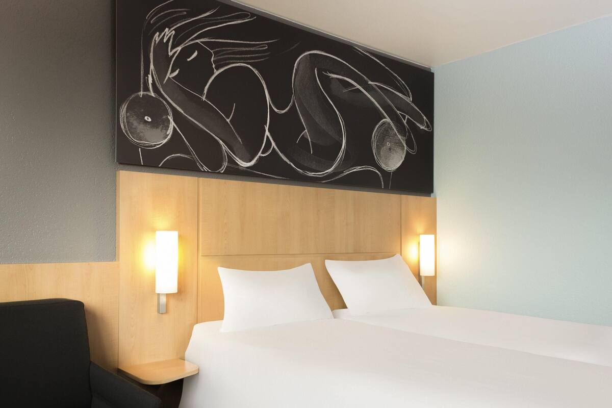 Quarto de hotel com uma cama, duas luminárias de cabeceira, um painel de cama de madeira e uma obra de arte na parede.