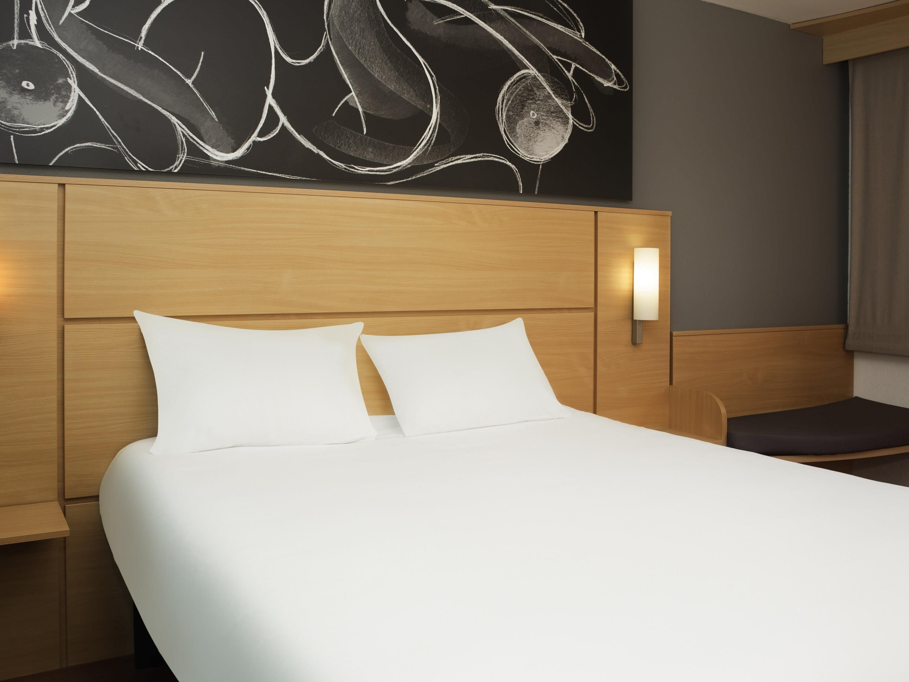 Ein Hotelzimmer mit einem hölzernen Kopfteil, einem Bett mit weißer Bettwäsche, einer Wandleuchte und einer Bank.