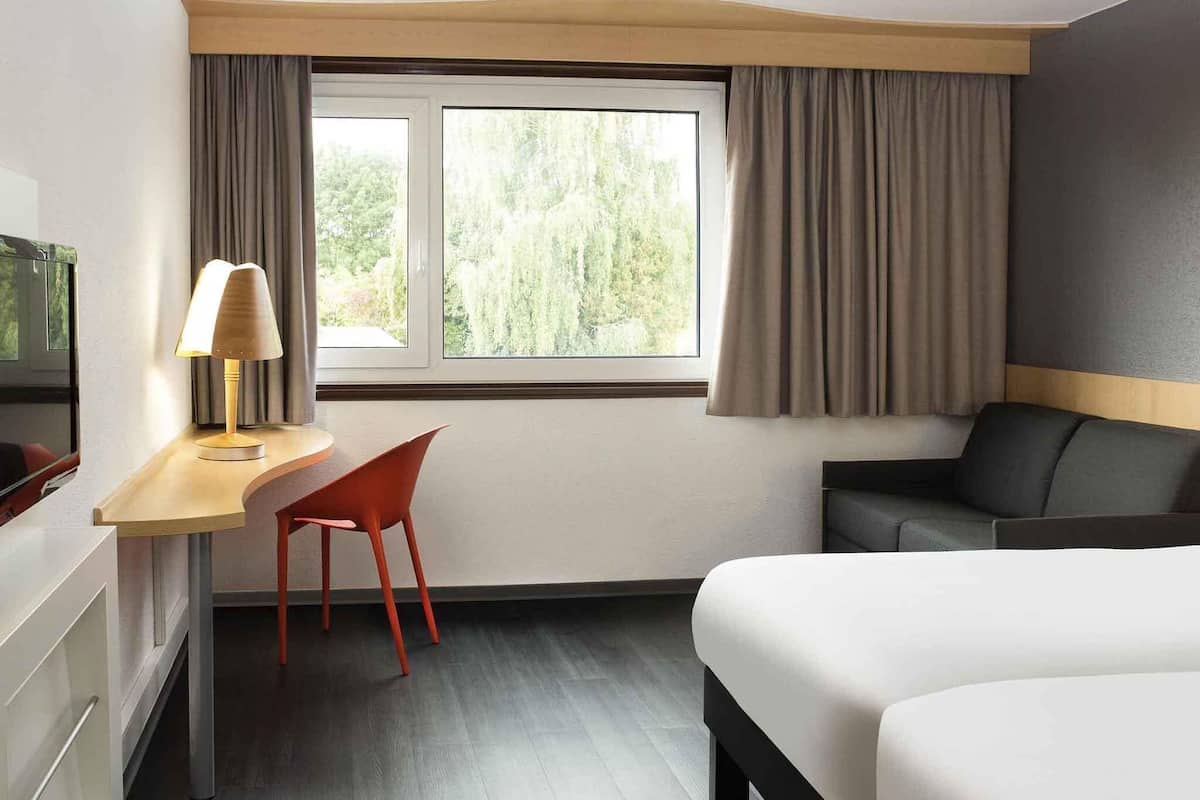 Ett hotellrum med en säng, ett skrivbord med en lampa, en stol, en tv och ett fönster med gardiner.