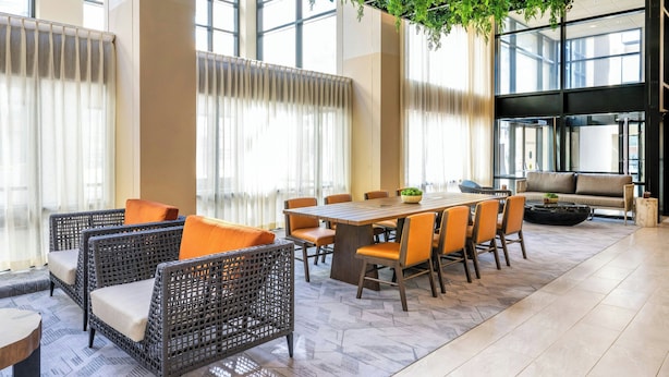 A modern hotel lobby with a large dining table, wicker chairs, a sofa, and a hanging green plant.