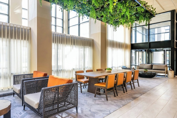 A modern hotel lobby with a large dining table, wicker chairs, a sofa, and a hanging green plant.