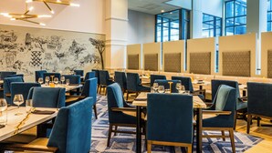 A modern dining area with blue upholstered chairs, white tables, and a large mural on the wall.