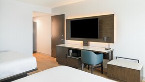 A modern hotel room with a bed, a desk with a chair, a television, and a minibar.
