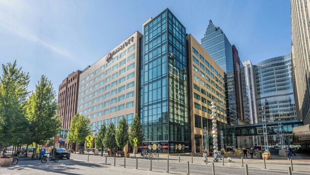 A Marriott hotel with a modern glass facade, surrounded by other tall buildings, trees, and a pedestrian area with benches and a bike rack.