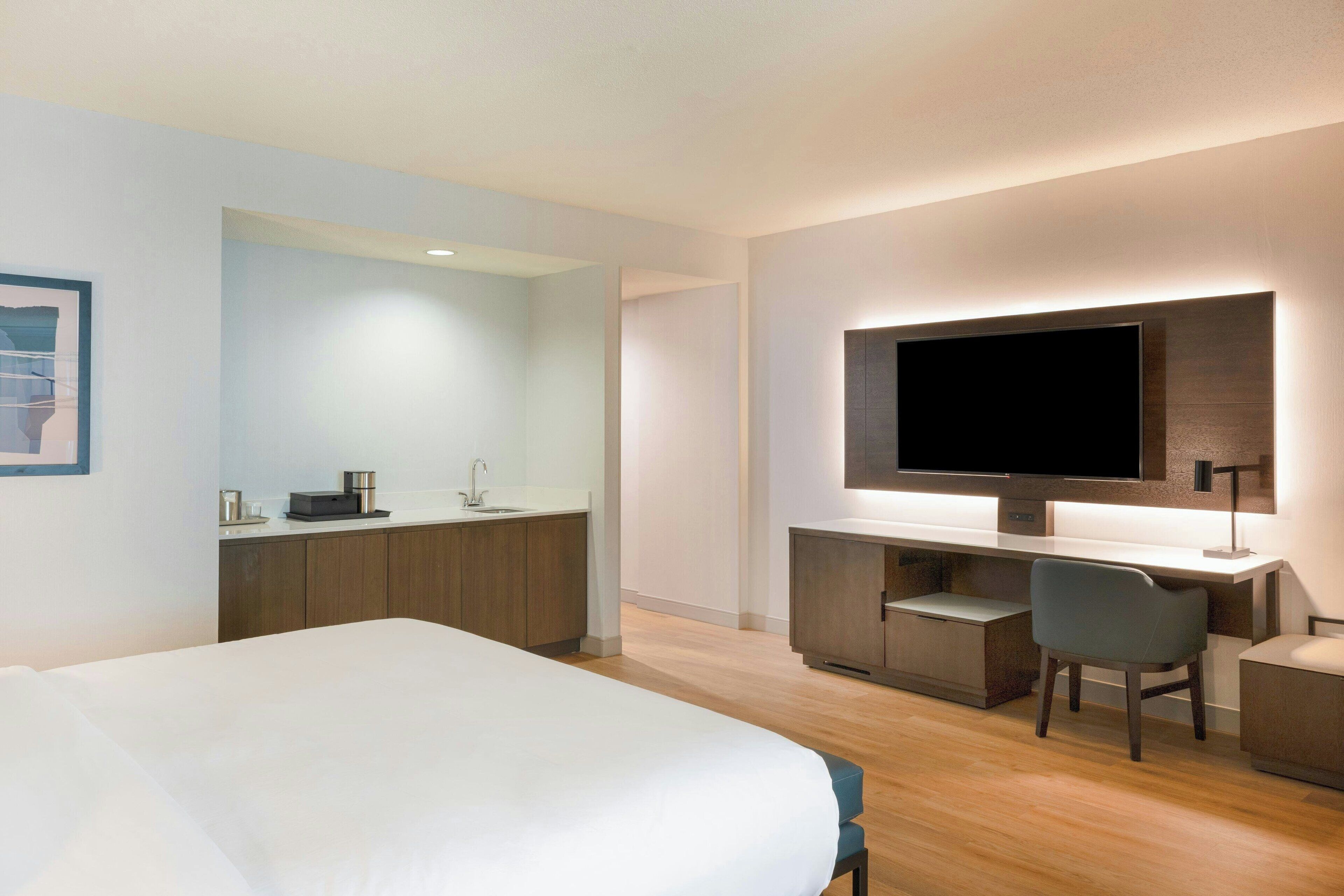 A modern hotel room with a large bed, a desk with a flat-screen TV, a chair, and a small kitchenette.