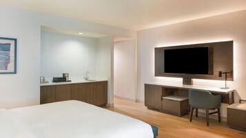 A modern hotel room with a large bed, a desk with a flat-screen TV, a chair, and a small kitchenette.
