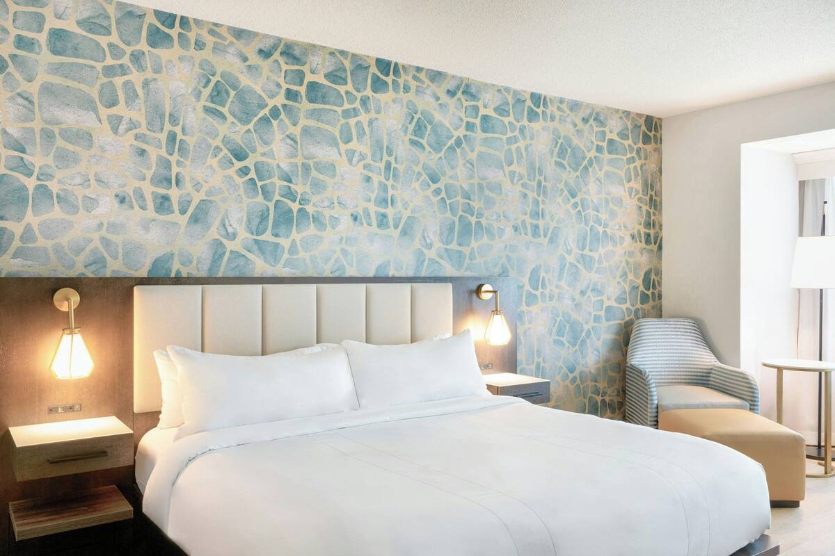 A hotel room with a large bed, a nightstand, a chair, and a stone-patterned wall.