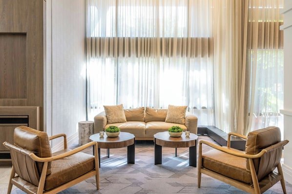 Una sala de estar moderna con un sofá de cuero color beige, dos mesas de centro de madera y un ventanal con cortinas traslúcidas.