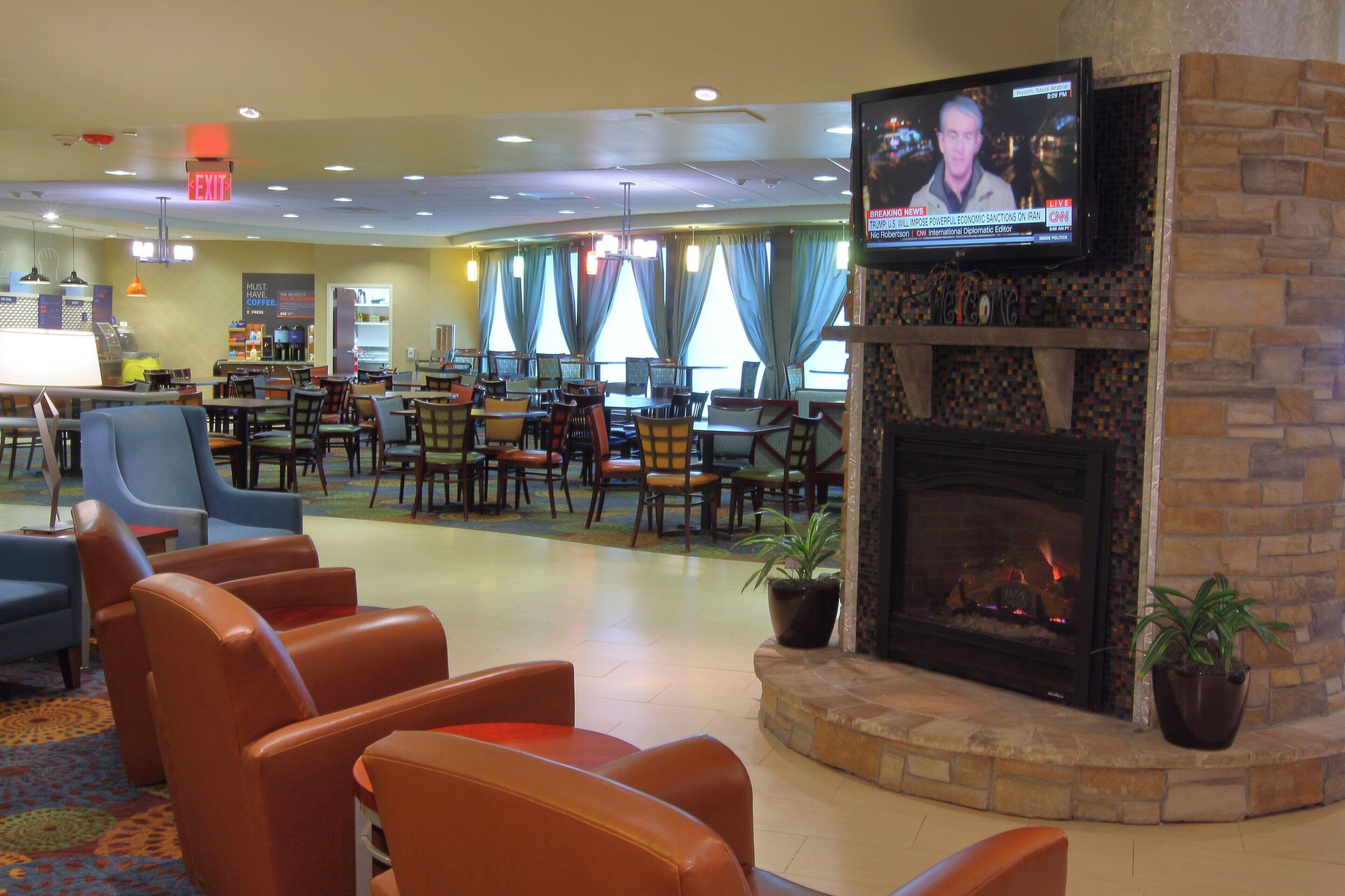 A hotel lobby with a television displaying a news program, seating areas with chairs and tables, a coffee station, and a fireplace.