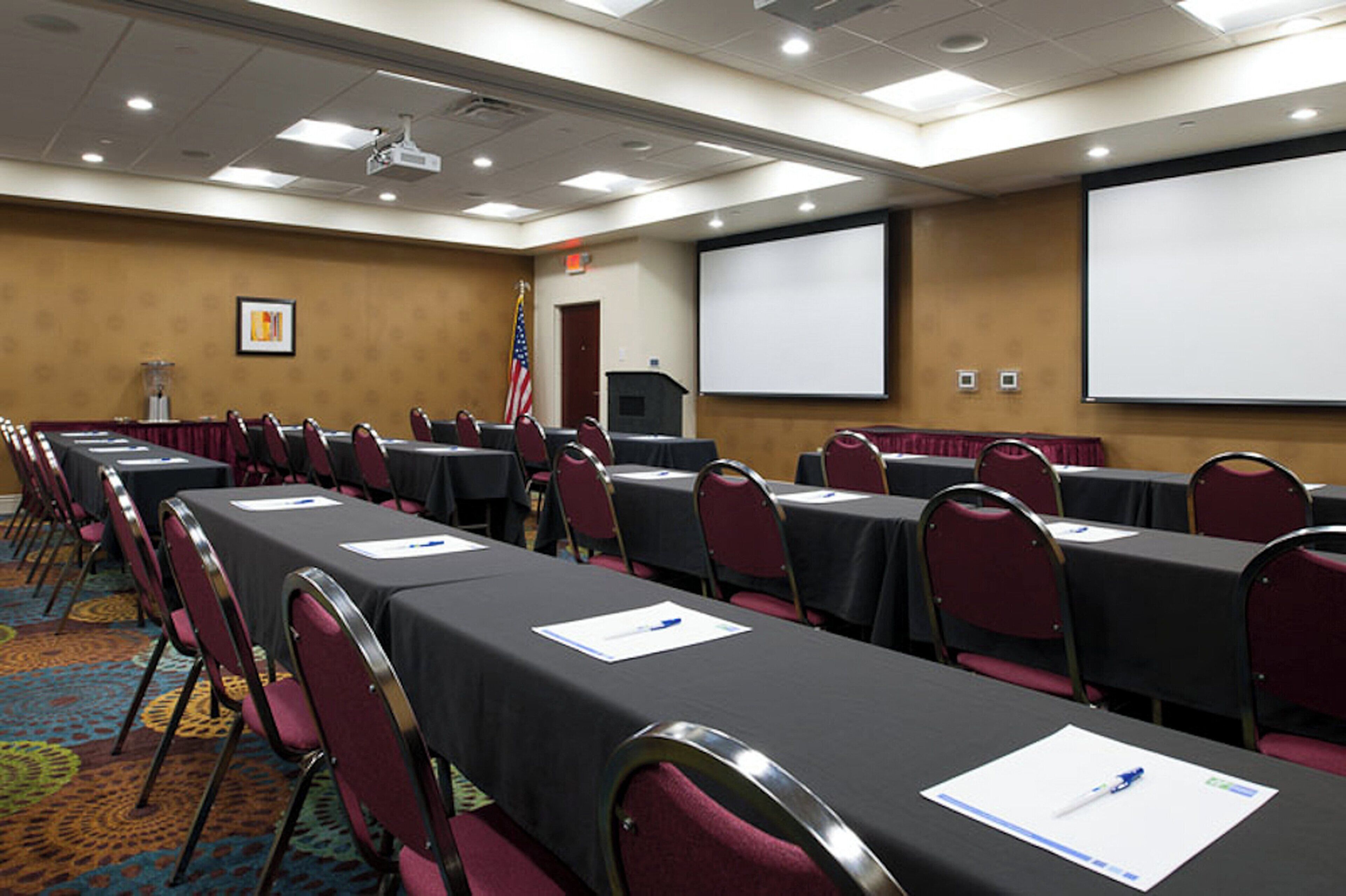 A conference room with rows of tables and chairs, a projector screen, and an American flag.