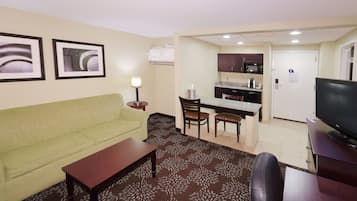 A hotel room with a green sofa, a wooden coffee table, a TV, a kitchen area with a microwave, and a patterned carpet.