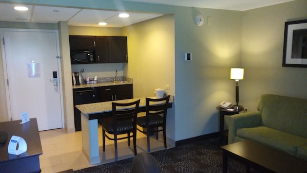 A compact hotel room with a kitchenette, a dining area with two chairs, a green sofa, and a small table.