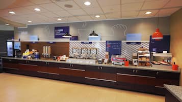 Espaço de buffet de café da manhã com diversas estações de comida, incluindo cereais, frutas, pratos quentes e produtos de panaria.