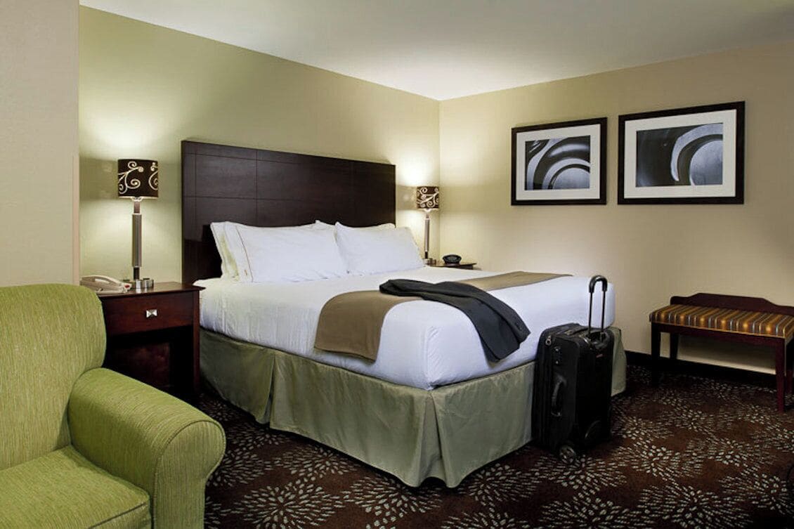 A hotel room with a bed, bedside lamps, a chair, a bench, and a suitcase.