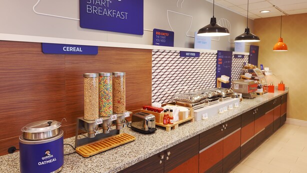 Espaço do buffet de café da manhã, com seções identificadas para cereais, comida quente e um dispensador de aveia Quaker.