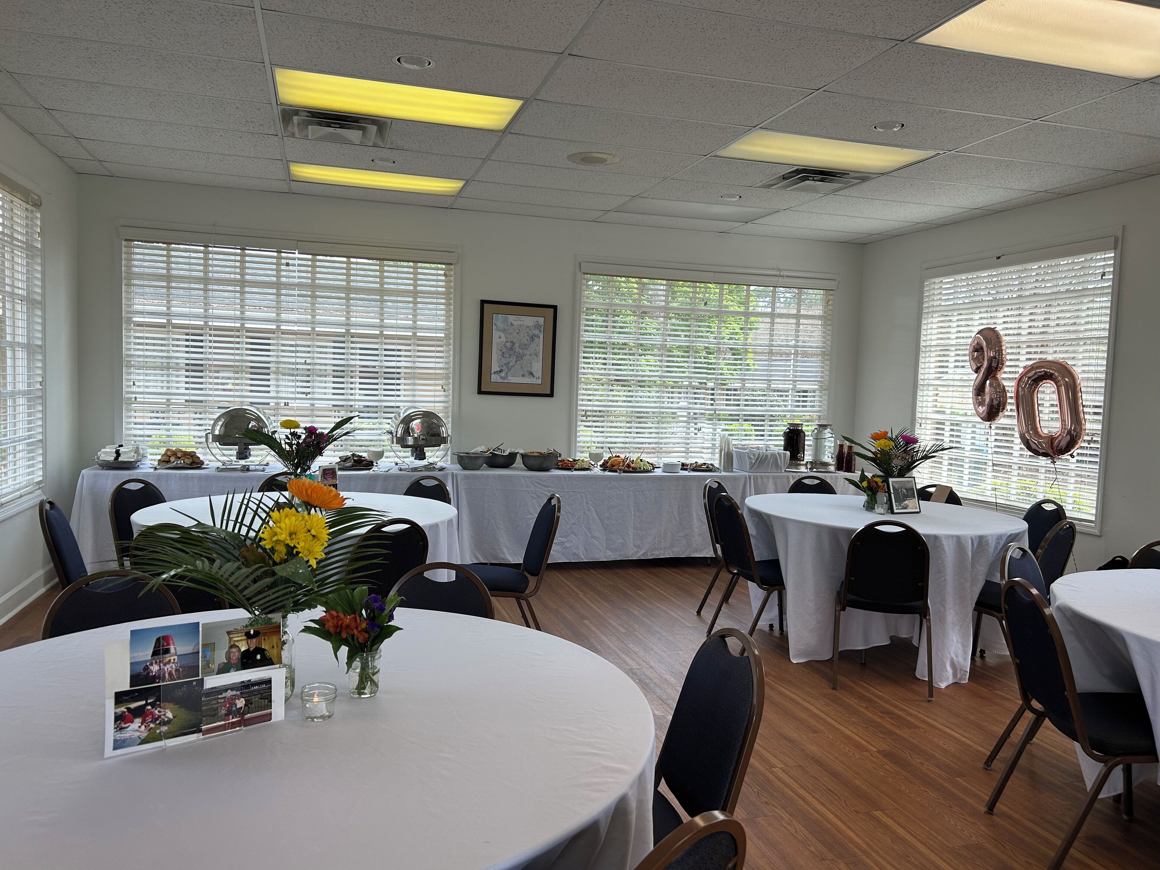 A dining area with round tables, white tablecloths, and chairs. There are windows with blinds, a framed picture on the wall, and a table with a floral arrangement.