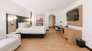 A modern hotel room with a bed, a desk, a chair, a TV, and a trash bin.