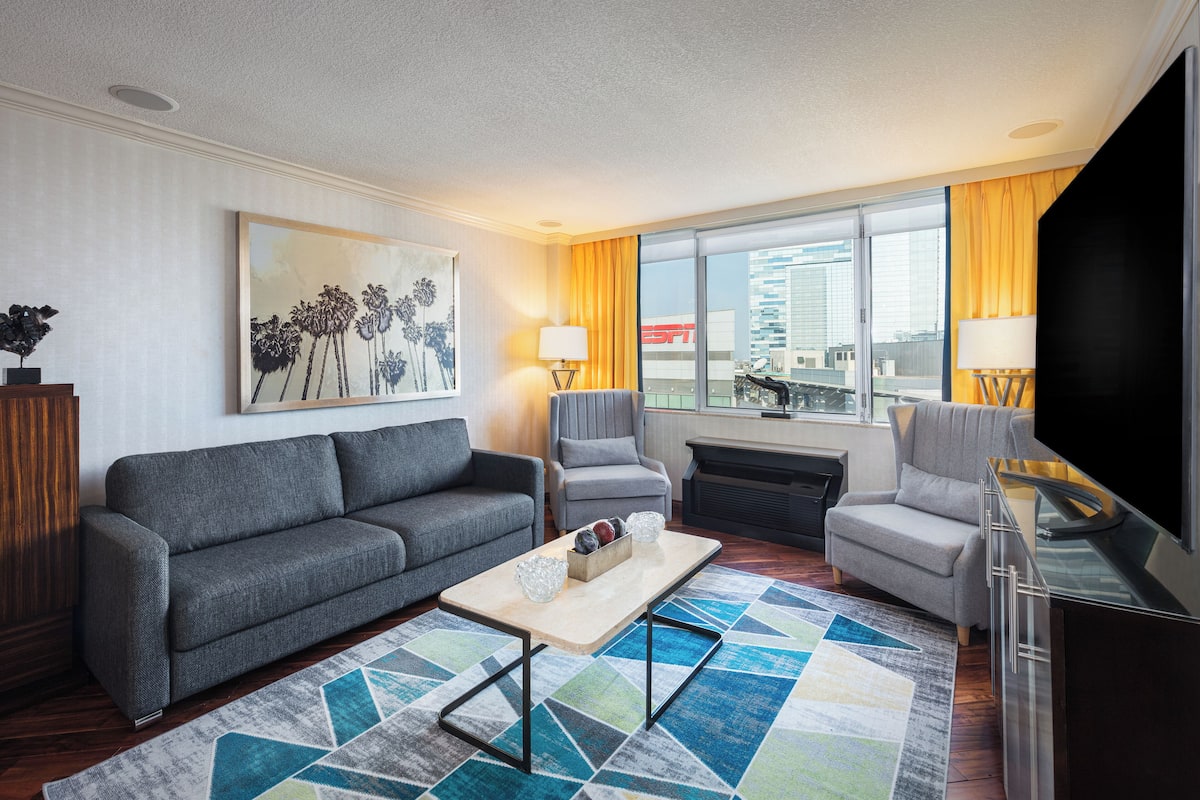 Une chambre d’hôtel moderne avec un canapé gris, deux fauteuils, une table basse et un téléviseur. La chambre est dotée de grandes fenêtres avec des rideaux, d’un tableau au mur et offre une vue sur un bâtiment à l’extérieur.