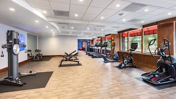 Uma academia com diversos equipamentos de exercício, piso de madeira e grandes janelas.