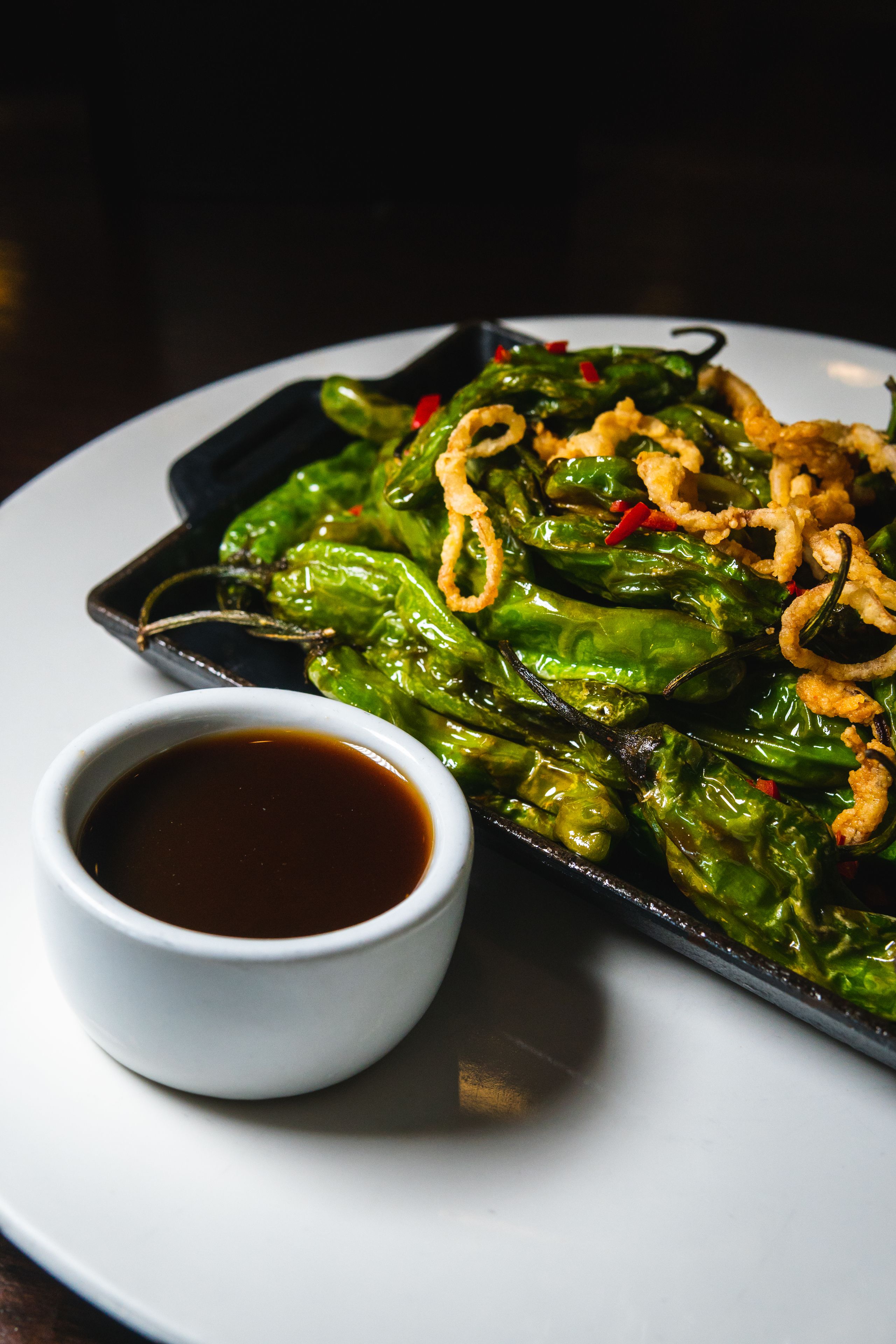 Un plat de poivrons verts grillés, accompagnés d’oignons frits et d’une petite portion de sauce.