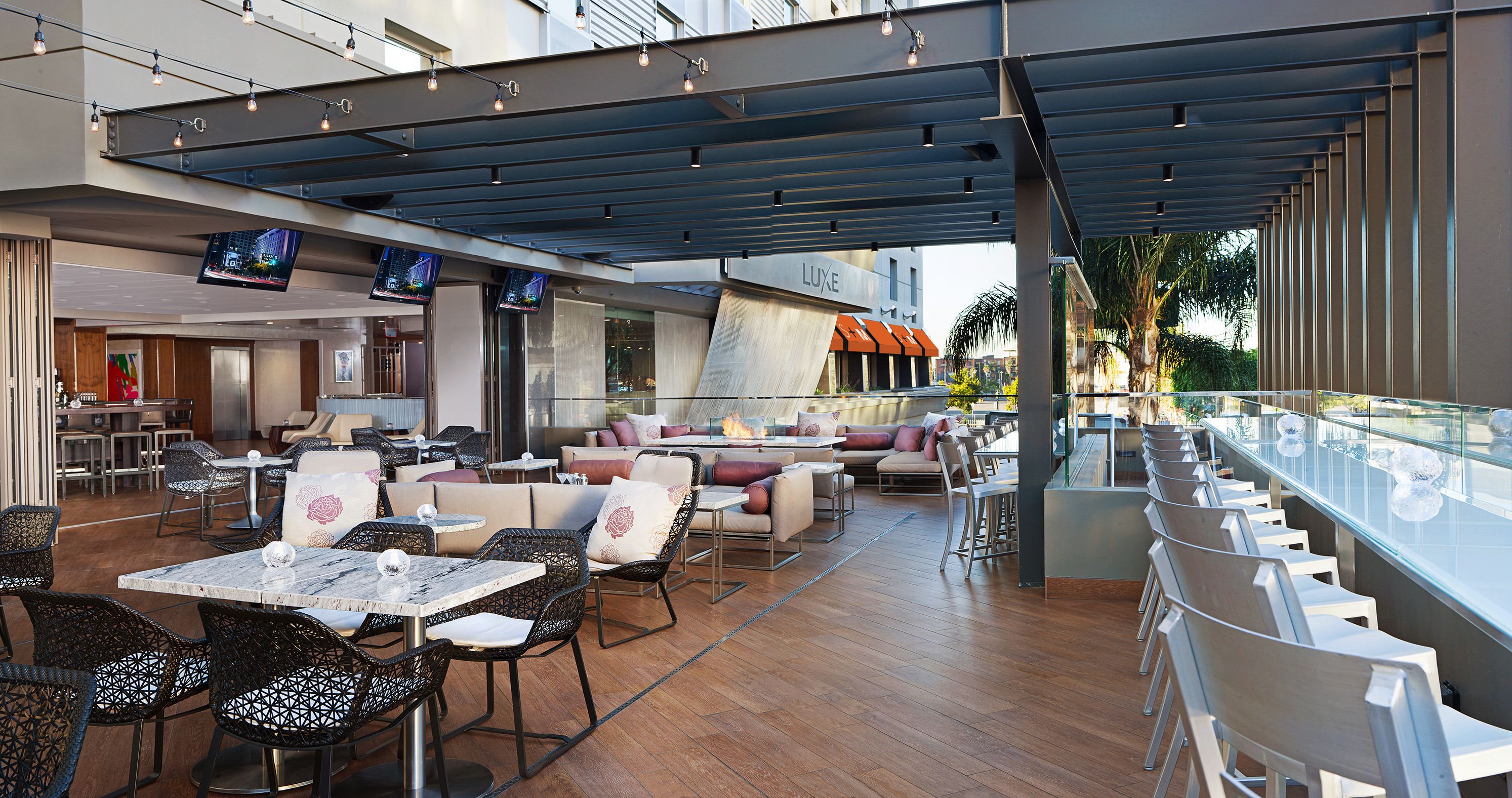 Un área de comedor exterior moderna con varias mesas y sillas, una barra con baranda de vidrio y vistas a un edificio con el nombre "LUXE".