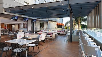Un área de comedor exterior moderna con varias mesas y sillas, una barra con baranda de vidrio y vistas a un edificio con el nombre "LUXE".