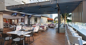 Un área de comedor exterior moderna con varias mesas y sillas, una barra con baranda de vidrio y vistas a un edificio con el nombre "LUXE".