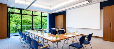 Un salón de conferencias con una larga mesa, sillas azules, una pantalla de proyector y grandes ventanales con vistas a zonas verdes.