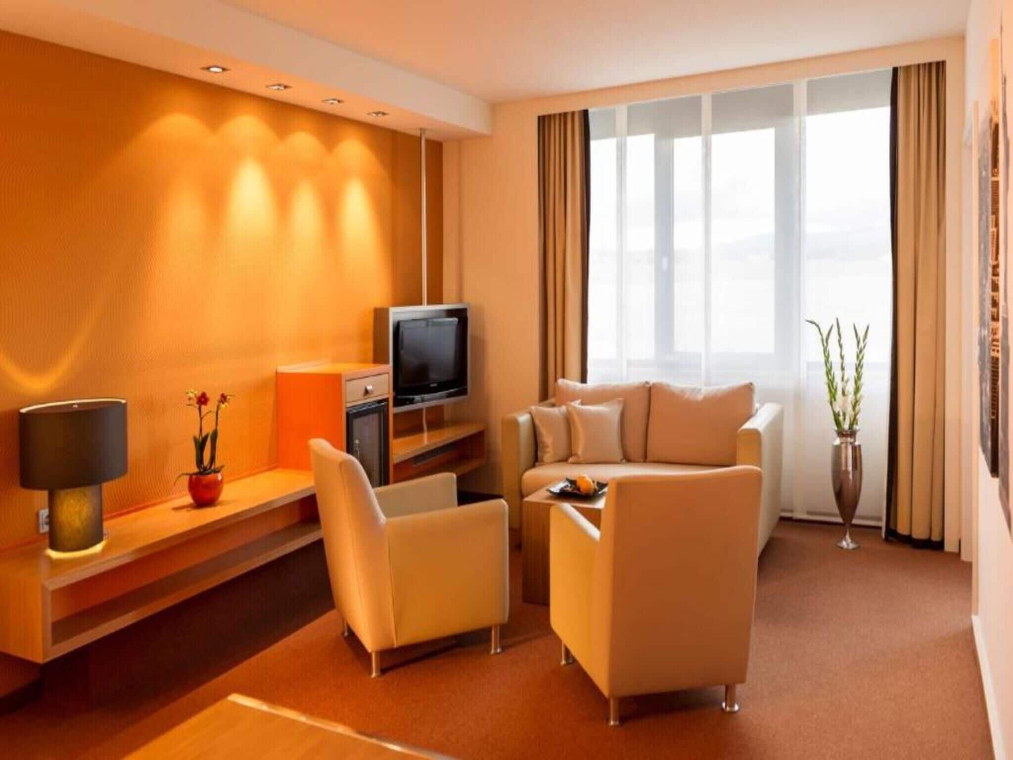 Une chambre d’hôtel avec un canapé, des fauteuils, une télévision, une lampe, une petite table et une plante.