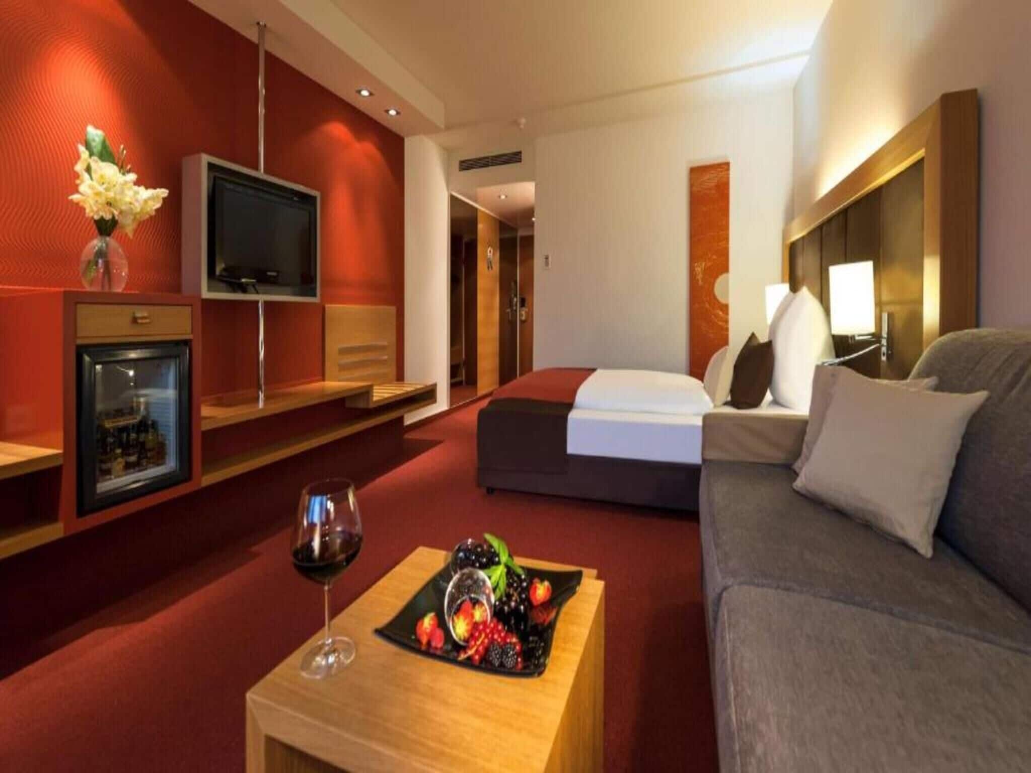 Hotellihuone, jossa on sänky, sohva, televisio, minibaari ja hedelmävati pöydällä.