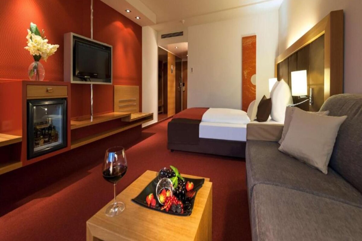 Ein Hotelzimmer mit Bett, Sofa, Fernseher, einer Minibar und einer Obstplatte auf einem Tisch.